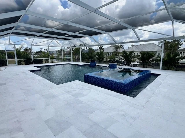 A modern L-shaped pool with a blue tiled raised spa under a large screened enclosure with travertine decking.
