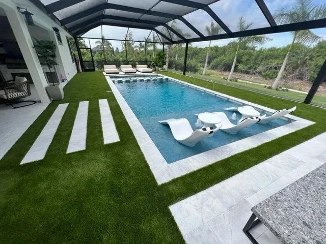 A rectangular pool with in-water loungers under a screened enclosure surrounded by artificial turf and white pavers.