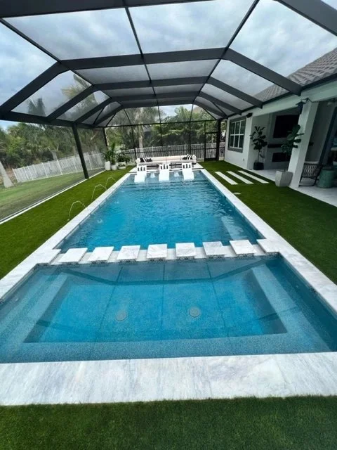 A screened rectangular pool with an attached raised spa, in-water loungers, and turf-lined deck under an overcast sky.