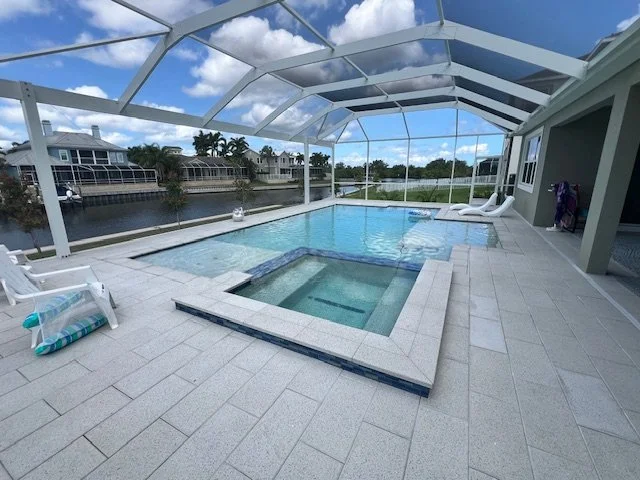 An L-shaped pool with an attached spa under a screened lanai overlooking a canal.