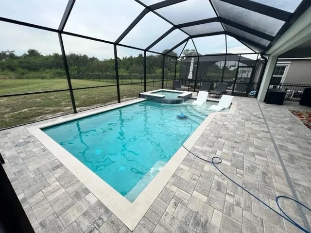 A rectangular pool with an attached raised spa under a screened lanai, surrounded by gray paver decking.