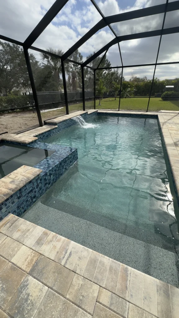 A small rectangular pool with a raised spa spilling water over a tiled wall, under a screened patio enclosure.