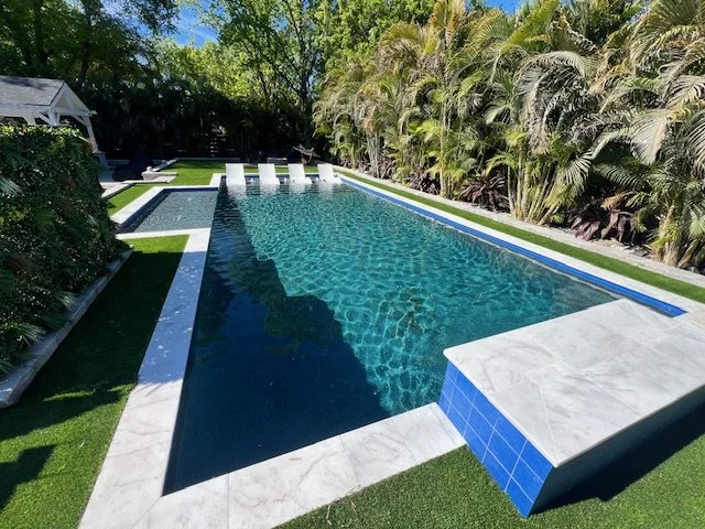 A rectangular pool with a tanning ledge and lounge chairs, surrounded by turf, palm trees, and a paver deck with a blue tile accent wall.
