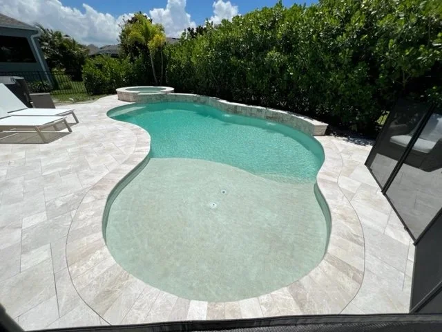 A freeform pool with a raised stone spa sits on a travertine paver patio under a screen enclosure.