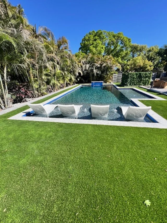 A rectangular pool with four white water-spouting scupper bowls set into a raised wall, surrounded by artificial turf and tropical landscaping.