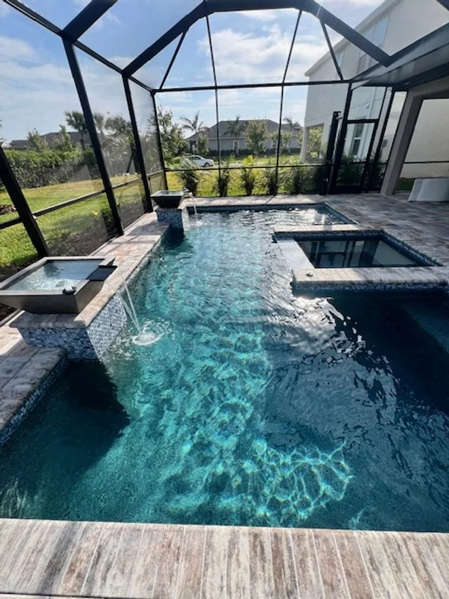 A geometric pool with a raised spa and two water-spilling bowls under a screened lanai.