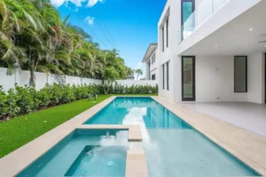 A modern lap pool with an integrated spa runs alongside a sleek white contemporary home framed by palm trees.
