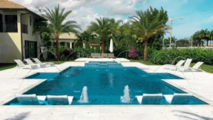 A rectangular backyard pool with deck jets arcing over in-pool loungers and a raised spa, surrounded by palm trees and white stone decking.