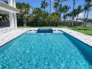 A rectangular backyard pool with a raised spa and arcing deck jets, framed by palm trees and a canal with a yacht in the background.
