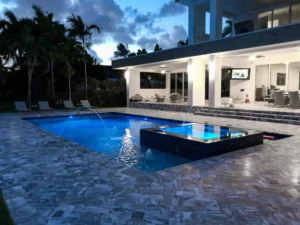 A rectangular pool with a raised glass-tile spa and dancing water jets glows blue at dusk beside a modern home.