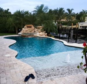 A freeform pool with a beach entry and rock waterfall spa, backing onto a canal with boats docked behind a black metal fence.