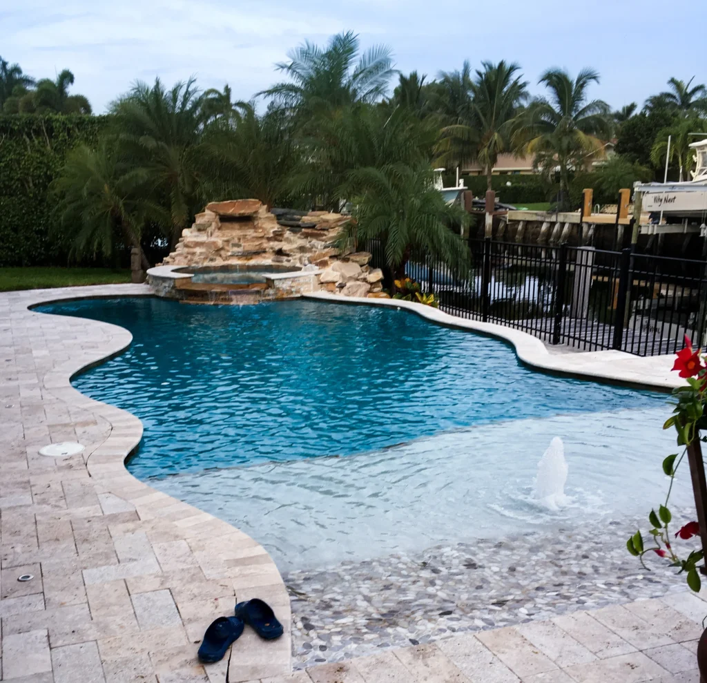 A freeform pool with a beach entry and rock waterfall spa, backing onto a canal with boats docked behind a black metal fence.