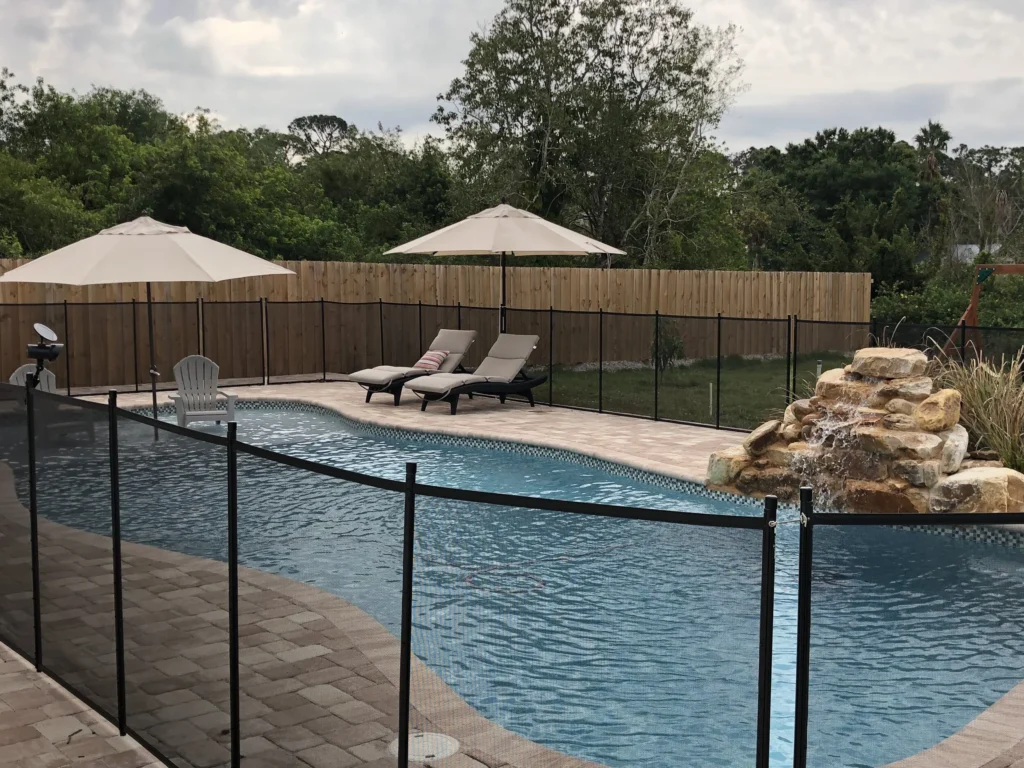 Freeform backyard pool with a rock waterfall feature, paver deck, loungers, and a removable child safety fence.