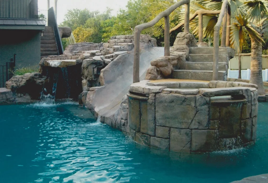 A rock-themed pool feature with a waterslide, waterfall, and raised stone spa surrounded by palm trees.