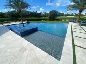 An infinity-edge pool with a dark tile raised spa overlooks a pond and golf course lined with palm trees.