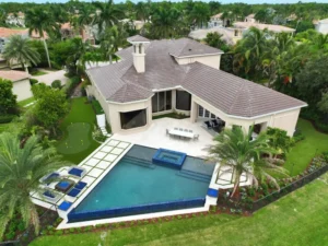 Aerial view of a geometric infinity-edge pool with raised spa beside a tile-roofed estate home and putting green.