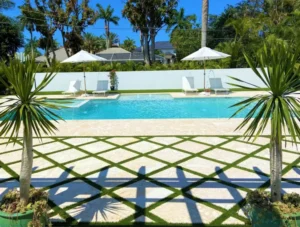 A rectangular backyard pool with water arcs, framed by palm trees, white lounge chairs, and a decorative turf-and-stone patio in the foreground.