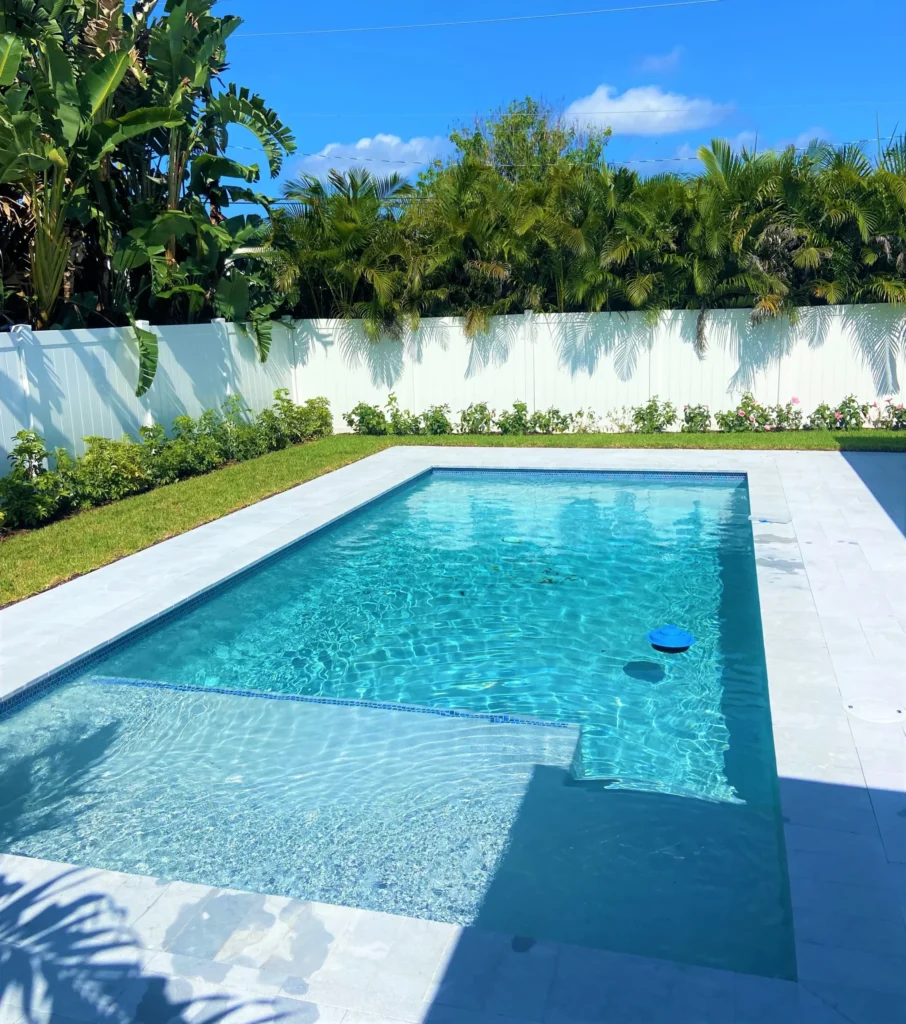 A small rectangular backyard pool with a shallow entry step, surrounded by travertine pavers and a white vinyl fence lined with tropical plants.