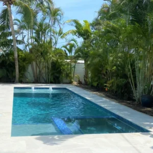 A rectangular backyard pool with two bubbler fountains, surrounded by tall palm trees and a travertine paver deck.