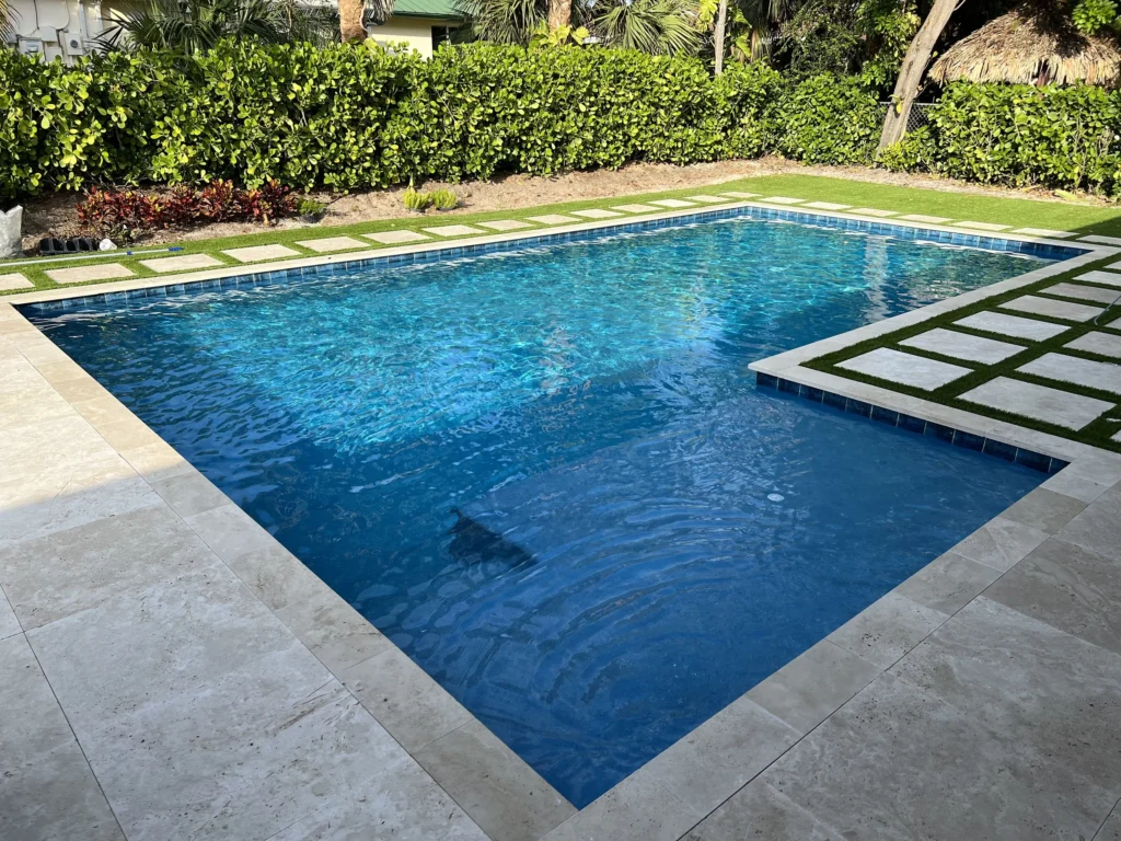 A rectangular backyard pool with blue tile waterline, travertine deck, and turf accents surrounded by hedges.