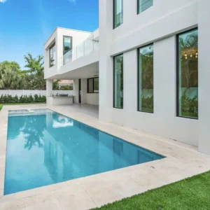 A long rectangular pool runs alongside a modern white two-story home with a raised spa at the far end.
