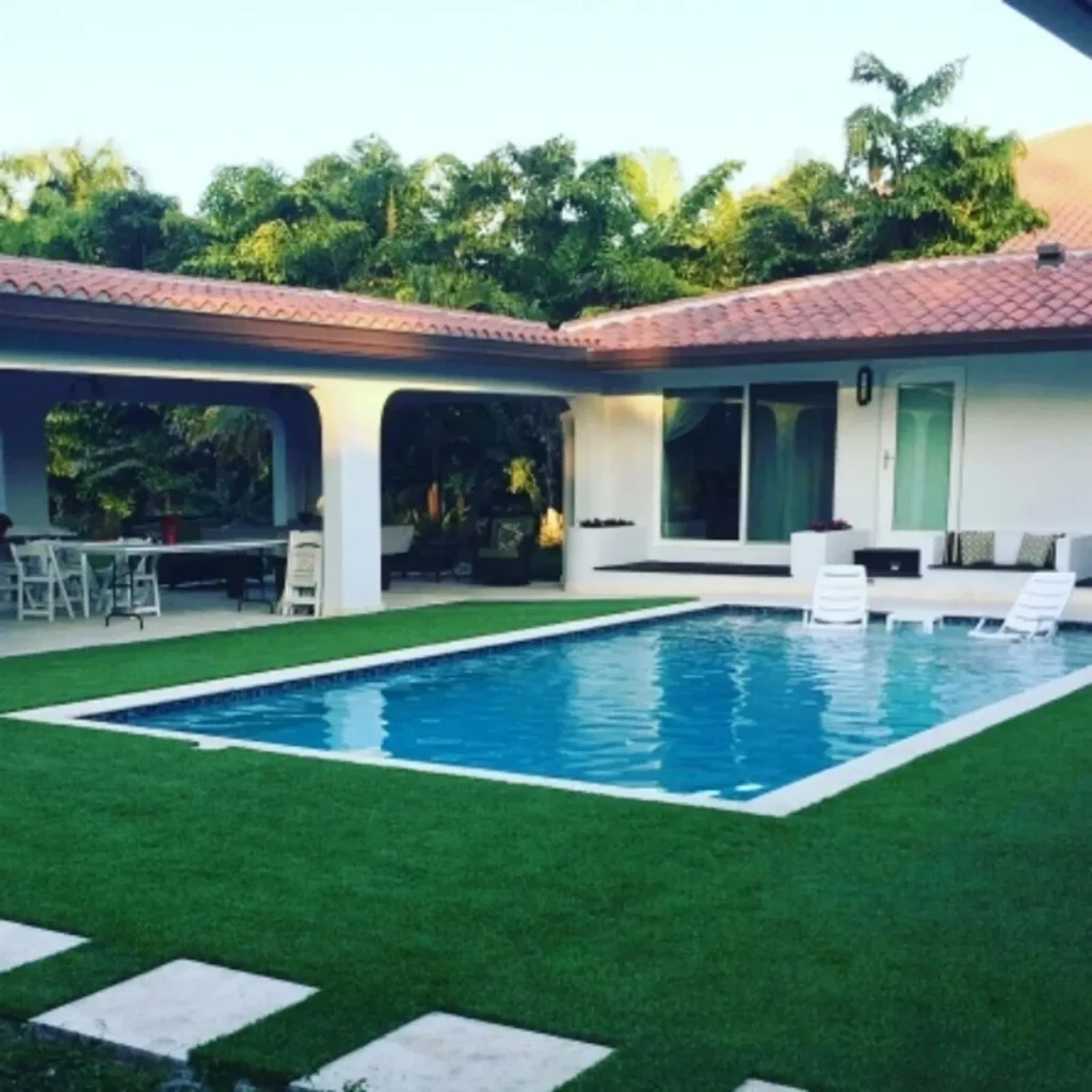 A small rectangular pool with turquoise water sits on a lush green artificial lawn beside a Spanish-tile-roofed home at dusk.
