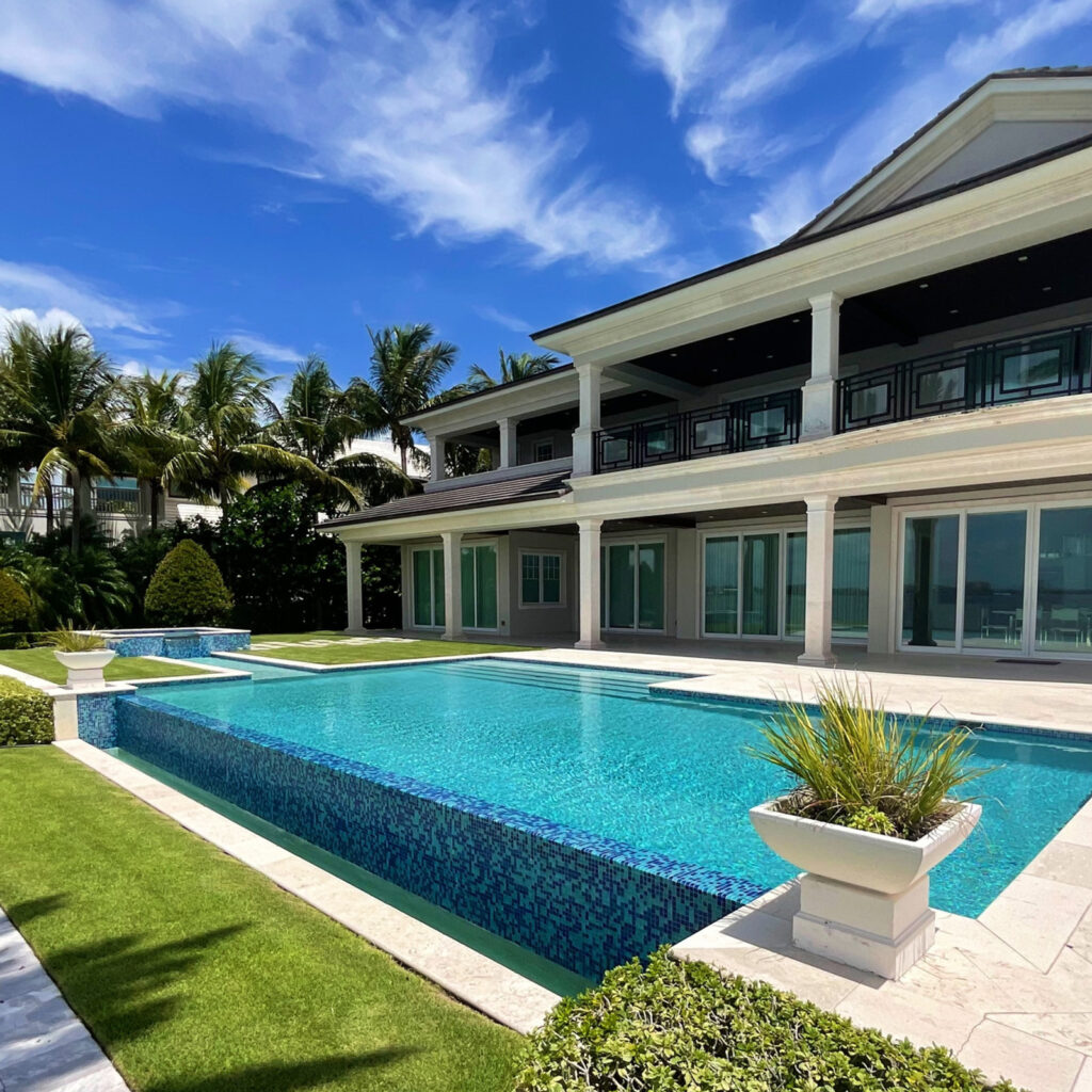 An elegant infinity-edge rectangular pool with blue mosaic tile walls and a raised spa beside a large modern home.