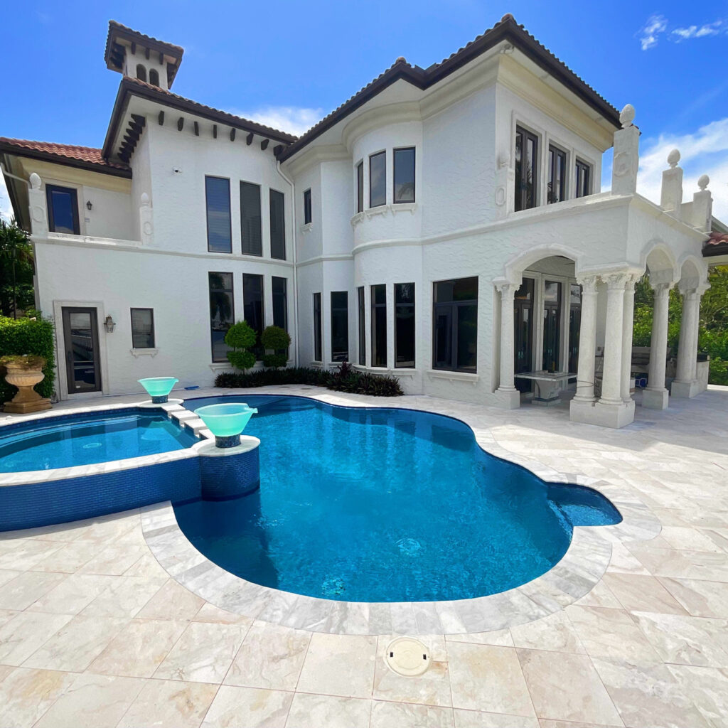 Large freeform pool with raised spa and glass water bowls in front of an elegant white stucco mansion with travertine deck.