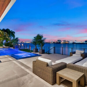 A waterfront infinity pool and raised spa glow at dusk beside a lounge deck overlooking a harbor.