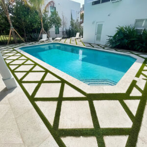 A geometric pool with clipped corners set in a decorative travertine and turf-lattice patio beside a white modern home.