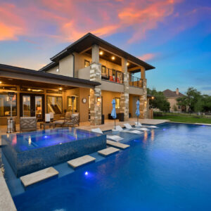 A large geometric infinity-edge pool with a raised glass-tile spa and floating loungers beside a two-story house at dusk.