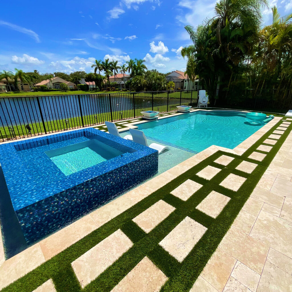 A rectangular backyard pool with a raised glass-tile spa beside a lake, framed by travertine deck and turf inlay.