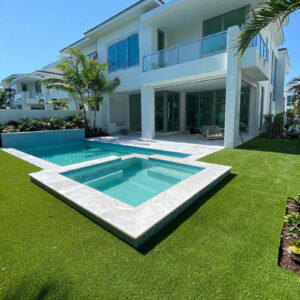 Modern rectangular pool with raised spa and blue glass tile accent wall behind a white two-story house with artificial turf yard.