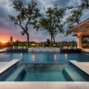 A raised spa with tiled steps sits in front of a pool and a stone wall with fire bowls and fountain jets at sunset.