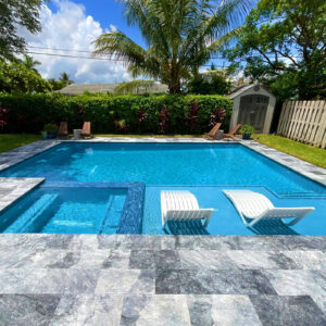 A rectangular backyard pool with a shallow sun shelf holding two white loungers, surrounded by gray stone pavers and tropical landscaping.