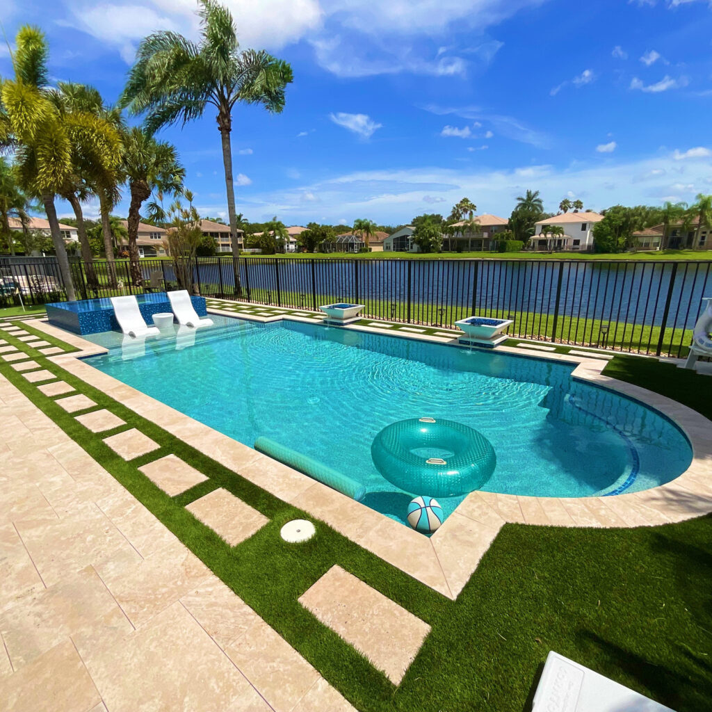 A freeform backyard pool with turquoise water, travertine decking, and a lake view behind a metal fence.