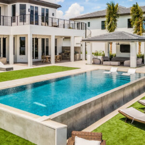 A raised rectangular infinity-edge style pool with in-water loungers sits between a modern two-story home and a covered cabana lounge.