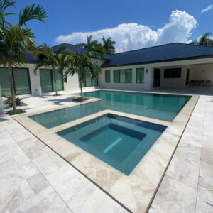 A geometric infinity-edge style pool with an integrated raised spa set into a travertine paver patio behind a modern single-story home.