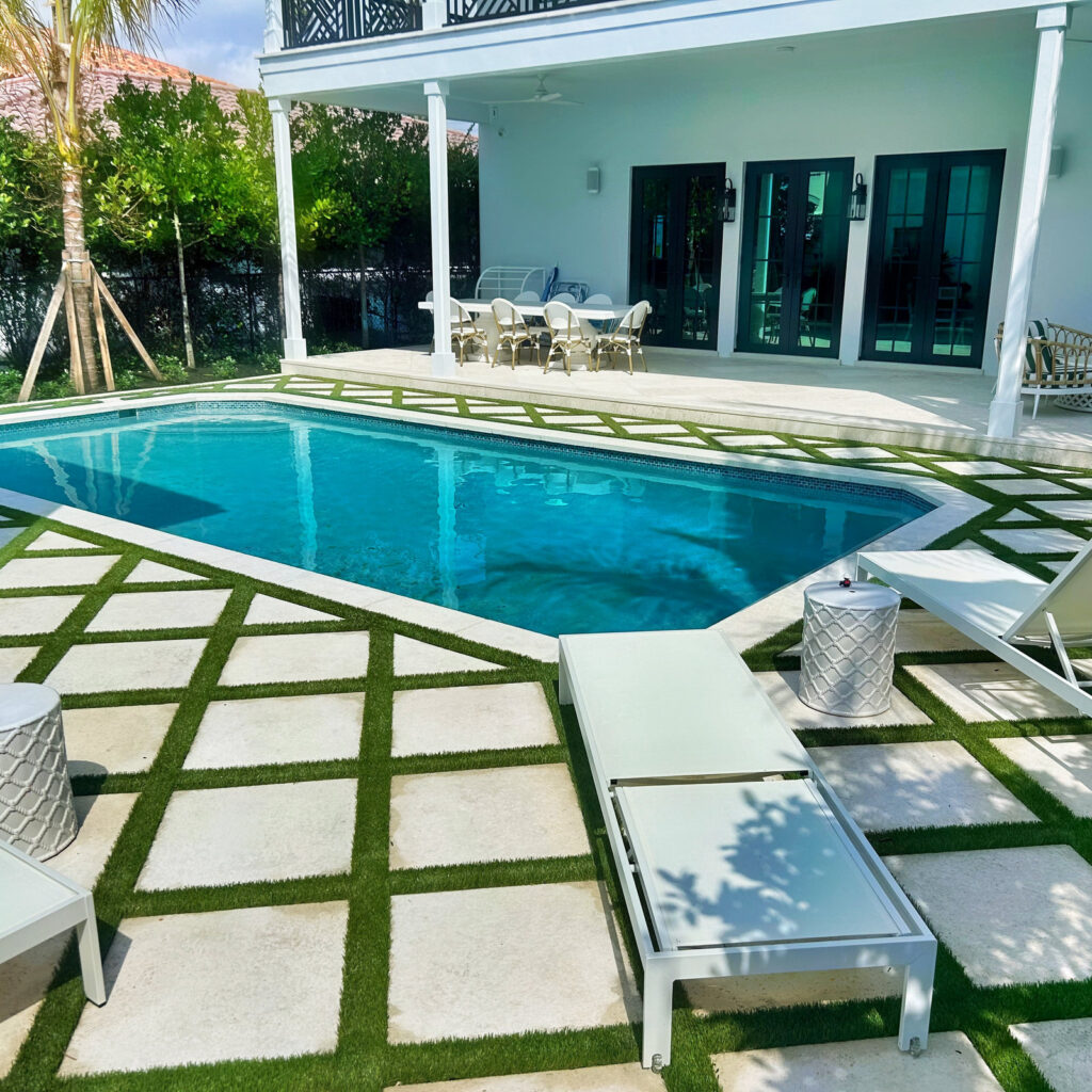 A rectangular backyard pool bordered by a checkerboard pattern of stone pavers and turf, with a covered patio and loungers nearby.