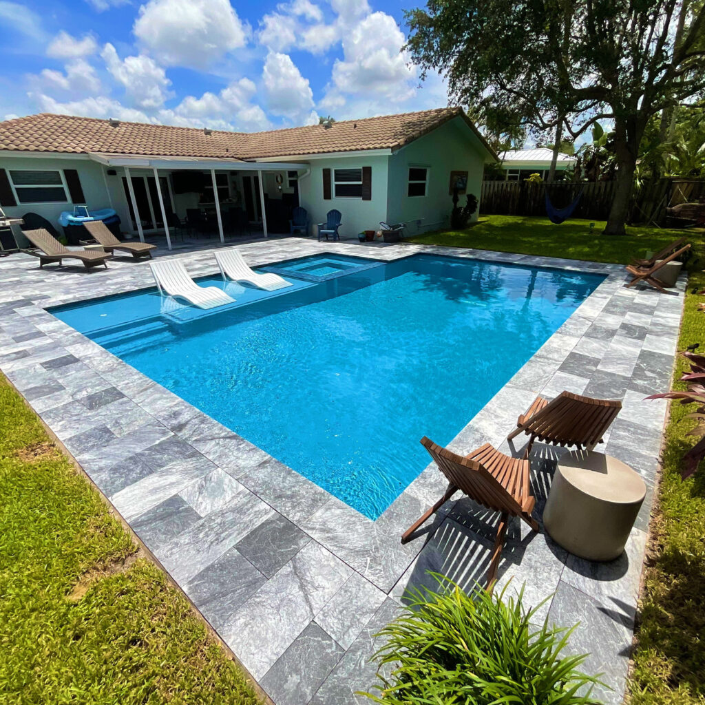 A small rectangular backyard pool with an attached spa, in-pool loungers, and a gray stone tile deck under open sky.