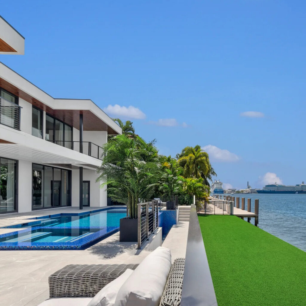 Modern waterfront home with an L-shaped pool along a paved terrace overlooking the bay.
