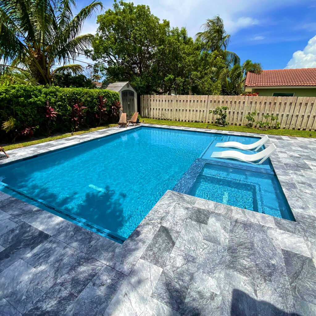 An L-shaped backyard pool with in-water loungers and a spa, surrounded by a gray stone patio and wooden fence.