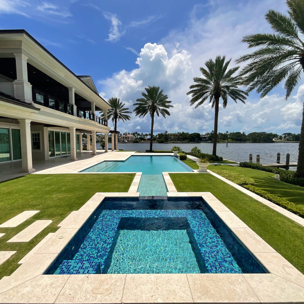 A tiled reflecting basin connects via a water channel to an infinity-edge pool overlooking a waterway, framed by palm trees.