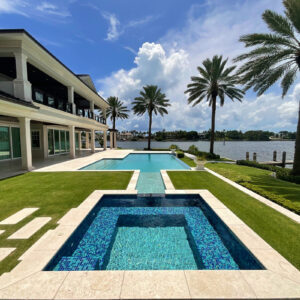 A tiled reflecting basin connects via a water channel to an infinity-edge pool overlooking a waterway, framed by palm trees.