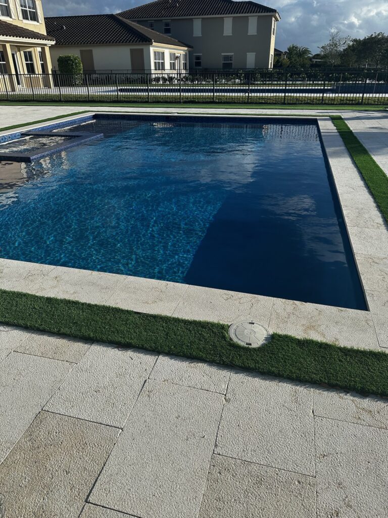 A rectangular pool with dark plaster and an attached raised spa on a travertine paver patio bordered by artificial turf.