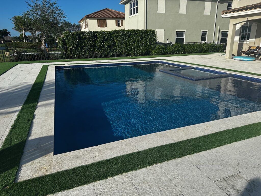 A rectangular pool with a dark interior and a flush-mounted spa, surrounded by paver decking and turf strips in a suburban backyard.