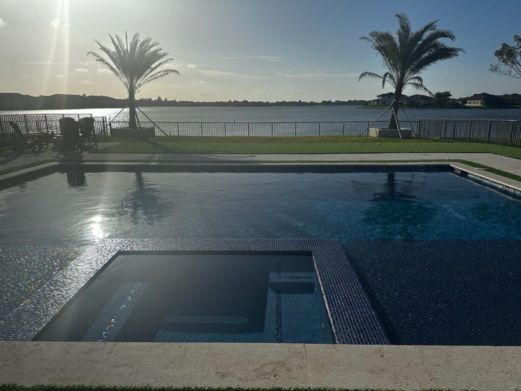 An infinity-edge style rectangular pool with dark glass tile spa overlooking a lake at sunrise.