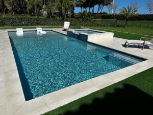 A rectangular pool with in-water loungers and a raised spa with a water feature, set on a travertine patio with turf lawn and palm trees.