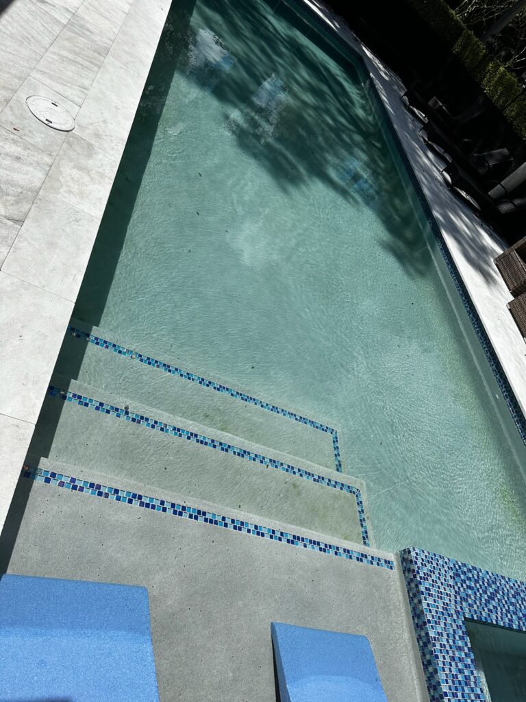 A steep overhead view of a pool corner with blue mosaic-tiled steps and gray paver decking.