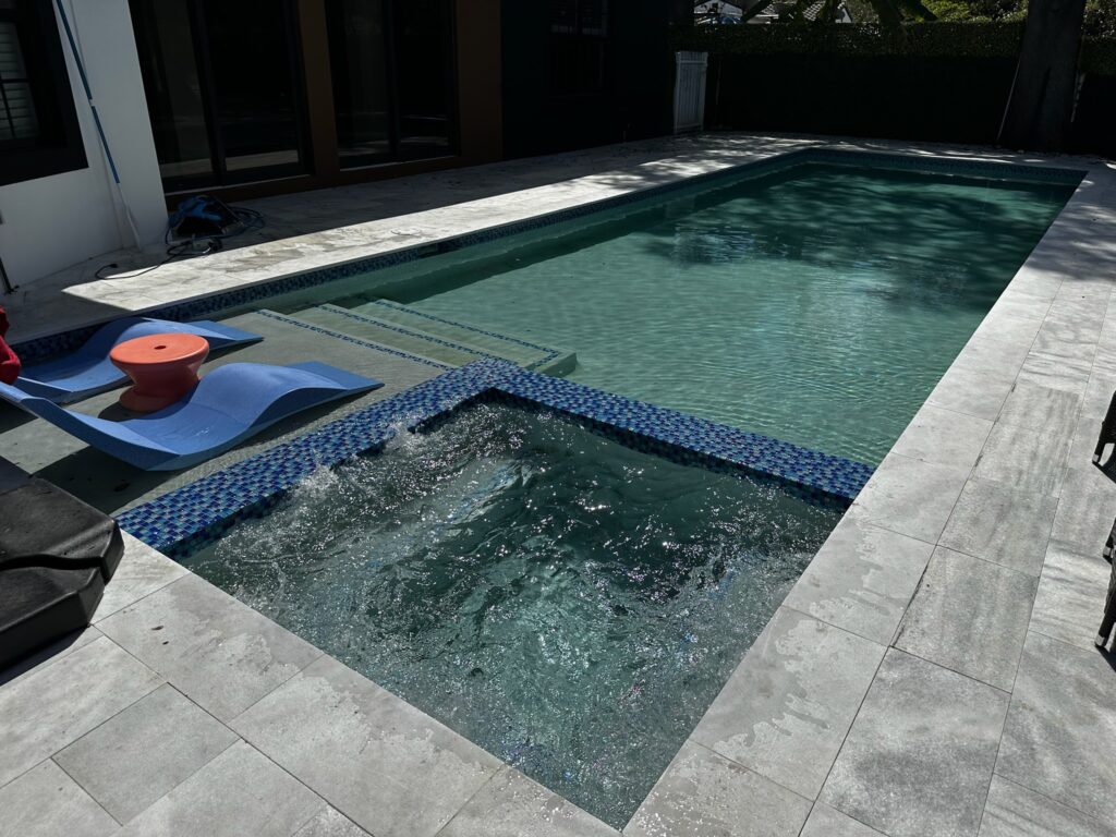 A rectangular pool with an integrated bubbling spa edged in blue mosaic tile and travertine deck.